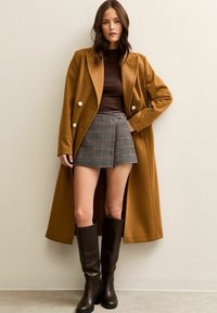 Vrouw in een mosterdkleurige lange jas, bruine top, houndstooth minirok en kniehoge donkerbruine laarzen, staande tegen een vlakke muur.