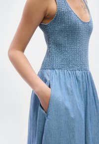 Μπλε φόρεμα από τζιν με κορσάζ με σφηκοφωλιά, στρογγυλή λαιμόκοψη και φούστα με πιέτες και πλαϊνές τσέπες. Λεπτή υφή και καθημερινός σχεδιασμός.