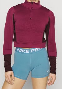 Camiseta atlética de manga larga de color granate con patrón texturizado y media cremallera, combinada con pantalones cortos azul claro con un logo de Nike blanco y banda.