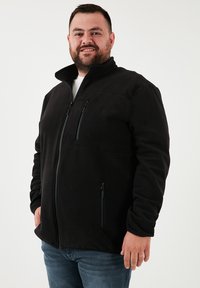 Chaqueta de forro polar negra con cuello alto, que cuenta con dos bolsillos en el pecho con cremallera y una cremallera frontal, llevada sobre una camisa blanca.