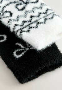 Kötött gyapjú csuklópántok, az egyik fekete, a másik fehér. Mindkettő kontrasztos geometriai mintákkal és puha, bolyhosszú textúrával rendelkezik.