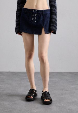Denim-Minirock mit Ösenakzenten und Schnürdetails vorne; kombiniert mit schwarzen Sandalen mit Riemchen auf einem strukturierten grauen Boden.