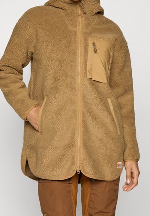 Braune Fleecejacke mit durchgehendem Reißverschluss, einer Reißverschlusstasche auf der Brust, Seitentaschen und abgerundetem Saum, getragen über brauner Hose.