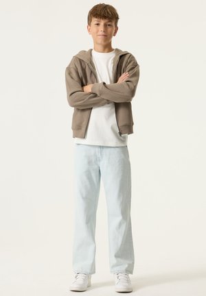 Hoodie marron à fermeture éclair porté sur un t-shirt blanc, associé à un jean bleu clair et des baskets blanches. Coupe décontractée avec un design confortable.