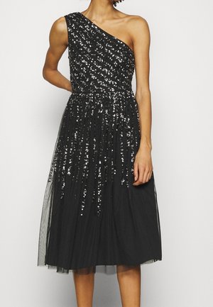 Czarna sukienka midi na jedno ramię z srebrnymi cekinami w ukośne pasy i pionowe wzory, połączona z przejrzystą spódnicą nałożoną na wierzch.