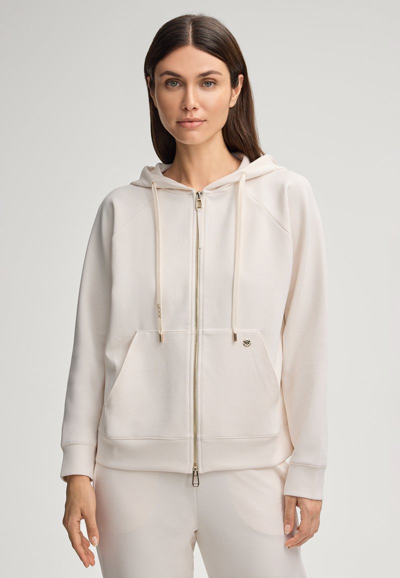 Hoodie zippé beige clair avec une poche kangourou, un capuche à cordon de serrage et des poignets côtelés. Il présente un détail de logo sur la poitrine gauche. Texture lisse.