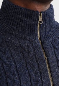 Marinblå kabelstickad tröja med hög krage, utrustad med en gyllene dragkedja. Texturen är tjock och fleece-liknande, med en något melerad uppsyn.