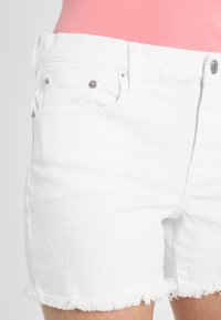 Pantalones cortos de mezclilla blancos con dobladillos deshilachados, que cuentan con herrajes plateados, bolsillos frontales y un diseño clásico de cinco bolsillos. Textura suave.
