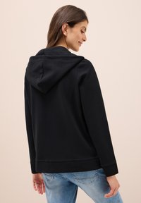 Felpa nera con cappuccio in tessuto morbido, dotata di cordino e polsini a costine. Indossata sopra jeans leggeri di colore azzurro con una vestibilità comoda.