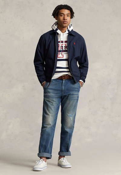 A young man stands confidently, wearing a navy jacket over a striped hoodie, paired with rolled-up jeans and white sneakers.