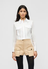 Νεαρή γυναίκα που φοράει λευκό πουκάμισο με μακριά μανίκια και κουμπιά και μπεζ σορτς ψηλόμεσο με μεγάλες μπροστινές τσέπες, συνδυασμένο με μαύρες καλσόν.