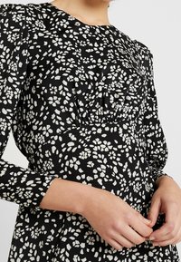 Svart klänning med vitt blommönster, långa puffärmar, rynkad frontdetalj och slät tygstruktur.