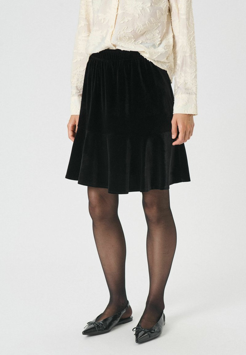 Zwarte fluwelen rok met een uitlopende snit en elastische tailleband. Model draagt zwarte flats en doorschijnende zwarte panty's. Textuur met een gladde afwerking.