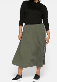 Jupe plissée verte olive avec détail de couture sur le côté, associée à un haut noir à manches longues et des baskets noires. Tissu doux, design jusqu'au genou.