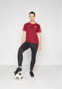 T-shirt de sport bordeaux avec des accents rouges et un logo, associé à un legging noir avec des rayures latérales rouges. Chaussures de football noires ; pied sur le ballon.