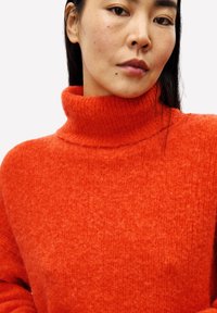 Vrouw met lang donker haar die een feloranje wollen coltrui draagt tegen een effen achtergrond.