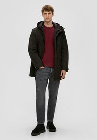 Schwarze Kapuzenparka mit glatter Oberfläche, kombiniert mit einem kastanienbraunen Pullover. Graue Jeans und schwarze Stiefel runden das Outfit ab, vor einem hellen Hintergrund.