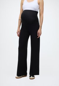 Raskaana oleva henkilö, jolla on mustat äitiyslahkeet, valkoinen toppi ja mustavalkoiset tennarit, seisoo käsivarret rentoina sivuilla.