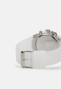 Correa de reloj de silicona blanca con hebilla de metal plateado, superficie texturizada y esfera de reloj circular con detalles mecánicos visibles en la parte posterior.