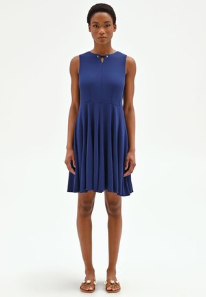 Donna in piedi che indossa un abito senza maniche blu navy con gonna plissettata e sandali beige decorati con grandi gemme decorative.