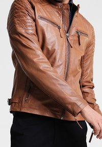 Giacca in pelle marrone con dettagli imbottiti sulle spalle, tasche anteriori con zip e design aderente. Texture liscia e linee eleganti.