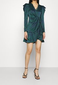 Zelené a námořnické modré zavinovací šaty se vzorem zebřích pruhů, dlouhými rukávy a volánkovým lemem. Kompletované s černými sandály na vysokém podpatku. Hladká textilie.