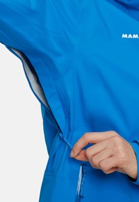 Giacca impermeabile blu con chiusura a cerniera laterale. Caratteristiche includono un cordino regolabile per una vestibilità ottimale, tessuto traspirante e superficie liscia.