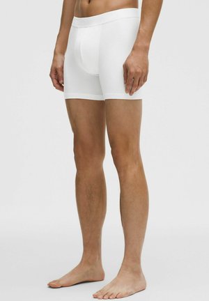 Uomo con boxer bianchi semplici, in piedi a piedi nudi contro uno sfondo chiaro neutro, gambe e parte bassa del busto visibili.
