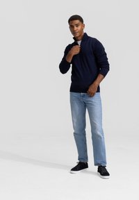 Navy pullover met een kwart-rits kraag, gecombineerd met lichtblauwe jeans en zwarte sneakers. Gladde textuur en slim-fit ontwerp.