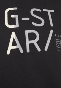 Tecido preto com letras grandes em cinza claro e branco formando "G-STAR/" e texto menor parcialmente visível no lado direito.