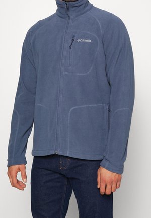 Mann trägt eine blaue Columbia-Fleecejacke mit Reißverschluss, Brust- und Seitentaschen, kombiniert mit dunkelblauen Jeans, steht vor einem schlichten Hintergrund.