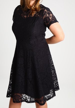 Frau trägt ein schwarzes Spitzenkurzarmkleid mit Blumenmustern, steht mit einer Hand hinter dem Rücken vor einem einfarbigen Hintergrund.