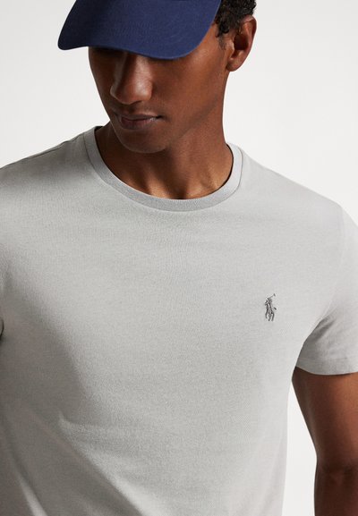 Γκρι βαμβακερό μπλουζάκι με στρογγυλή λαιμόκοψη και κοντά μανίκια, με μικρό κεντημένο λογότυπο στο στήθος. Το ύφασμα έχει απαλή υφή.