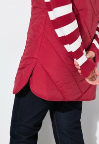 Chaleco acolchado rojo con un dobladillo asimétrico y costuras en ángulo, combinado con una camisa de rayas rojas y blancas de manga larga. Unos jeans oscuros completan el conjunto.