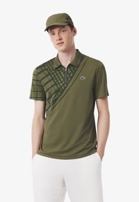 Olijfgroene polo met geometrisch patroon in donkergroen, korte mouwen, ritssluiting en Lacoste-logo op de borst. Vergezeld van een bijpassende pet.
