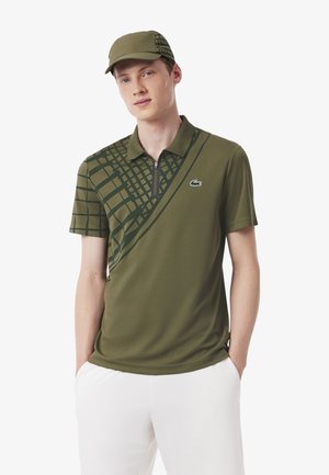Olijfgroene polo met geometrisch patroon in donkergroen, korte mouwen, ritssluiting en Lacoste-logo op de borst. Vergezeld van een bijpassende pet.