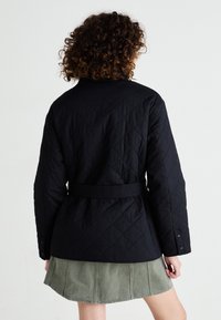 Jachetă cu quilted neagră, cu talie evidențiată de o curea, având un model texturat și o croială ajustată, asociată cu o fustă verde deschis.