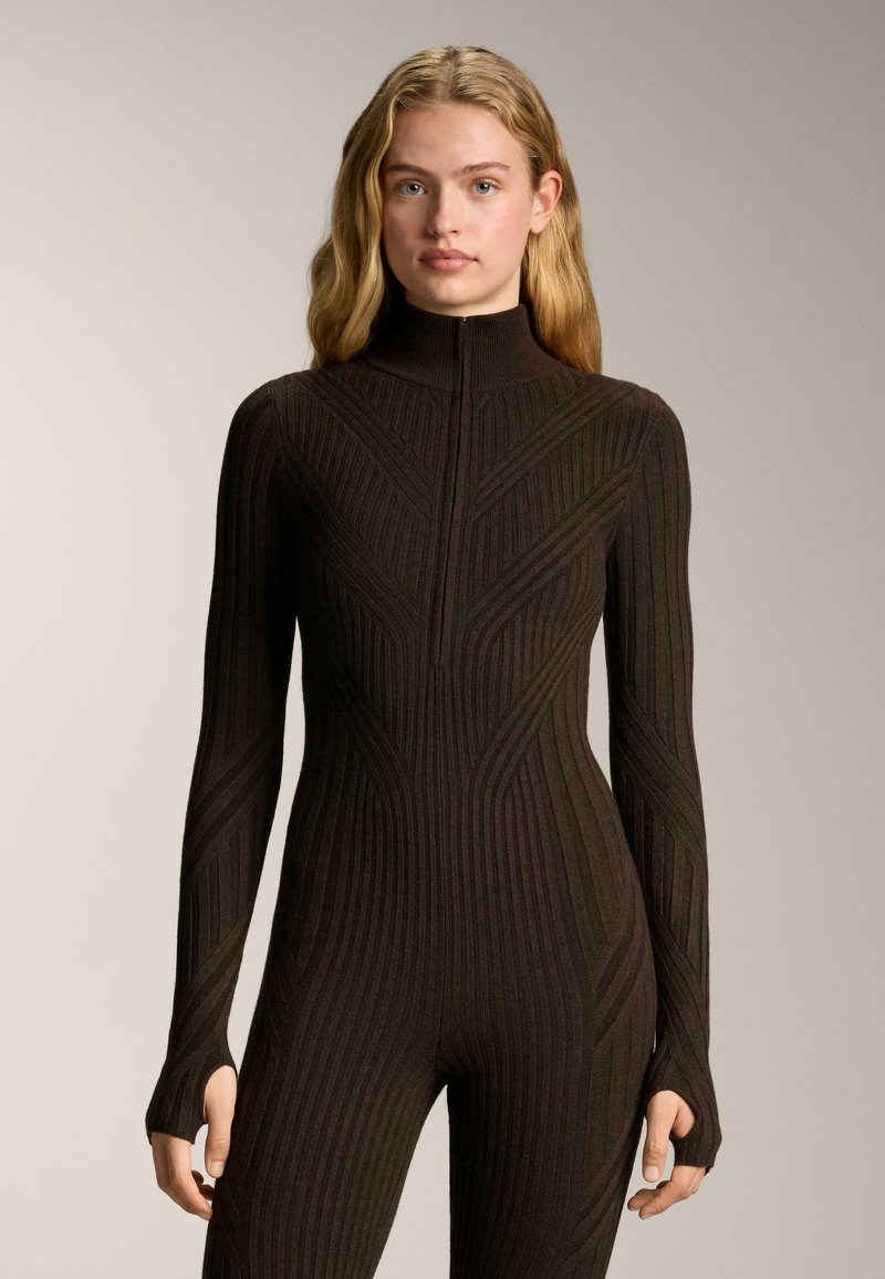 Žena plave kose nosi uski tamnobijeli kombinirani kombinezon rebraste teksture s dugim rukavima, visokim ovratnikom i otvorima za palce.