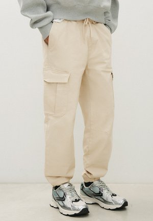Pantalon cargo beige avec des poignets élastiques porté avec des baskets athlétiques argentées et noires et un sweat gris. Mains dans les poches.