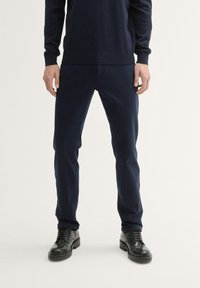 Pantalon bleu marine au design ajusté, avec une texture lisse et une finition subtile. Associé à des chaussures en cuir noires.