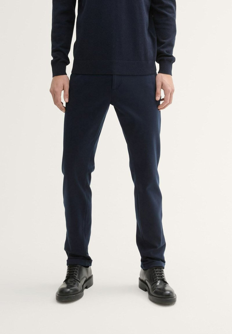 Pantalon bleu marine au design ajusté, avec une texture lisse et une finition subtile. Associé à des chaussures en cuir noires.