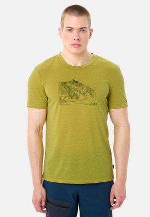 Giovane uomo con indosso una maglietta verde oliva con grafica di un paesaggio montano e pantaloni blu scuro, in piedi davanti a uno sfondo semplice.