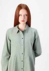 Abito camicia verde chiaro con maniche lunghe, apertura frontale con bottoni, colletto a punta e parte inferiore plissettata. Presenta bottoni scuri lungo la placchetta.
