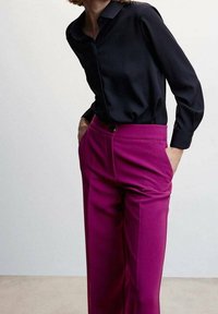Zwarte lange mouwen shirt met een kraag die opknopen kan, gecombineerd met hoge fuchsia broeken met een enkele knoop en zijzakken.