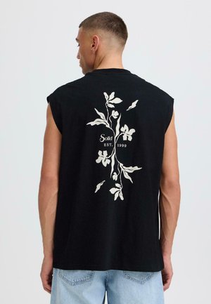 Homme portant un débardeur noir avec un motif floral blanc et le texte "Solid EST. 1999" au dos, associé à un jean bleu clair.