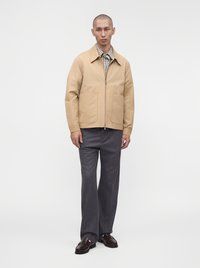 Homme portant une veste beige zippée, une chemise à carreaux, un pantalon gris et des chaussures marron foncé, debout devant un fond blanc uni.