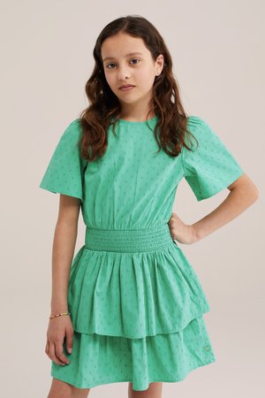 Ragazza con lunghi capelli castani indossa un vestito verde a maniche corte con vita arricciata e gonna a più strati, in piedi con una mano sul fianco.