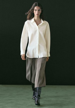 Donna che cammina su pavimento verde indossando una camicia bianca oversize con bottoni, pantaloni beige larghi e corti, e stivali neri al ginocchio su sfondo scuro.