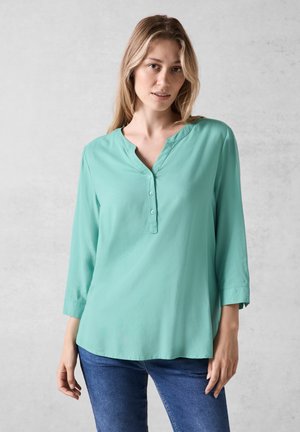 Blusa ligera en color aguamarina con escote en V, mangas de tres cuartos y acentos de botones. Textura suave con un corte holgado, combinada con vaqueros.