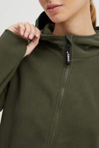 Giacca in pile verde oliva con zip frontale, cappuccio e tessuto testurizzato. Caratterizzata da un tirazip nero con dettaglio del logo.